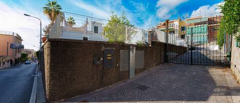 A cobblestone street with a brick wall and metal gate, leading to a building with a balcony and a palm tree.
