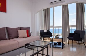 Ein modernes Wohnzimmer mit einer Couch, zwei Sesseln, einem Glastisch und Meerblick durch große Fenster.