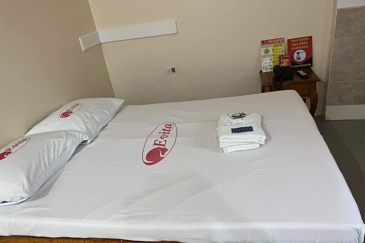 Et hotelværelse med en pænt redt seng, en pude med hotellets logo, et foldet håndklæde og et skilt, der angiver, at værelset er blevet rengjort.