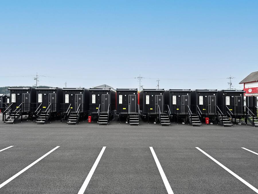 Una hilera de baños portátiles negros con números en las puertas, ubicados en un estacionamiento con líneas blancas en el suelo.