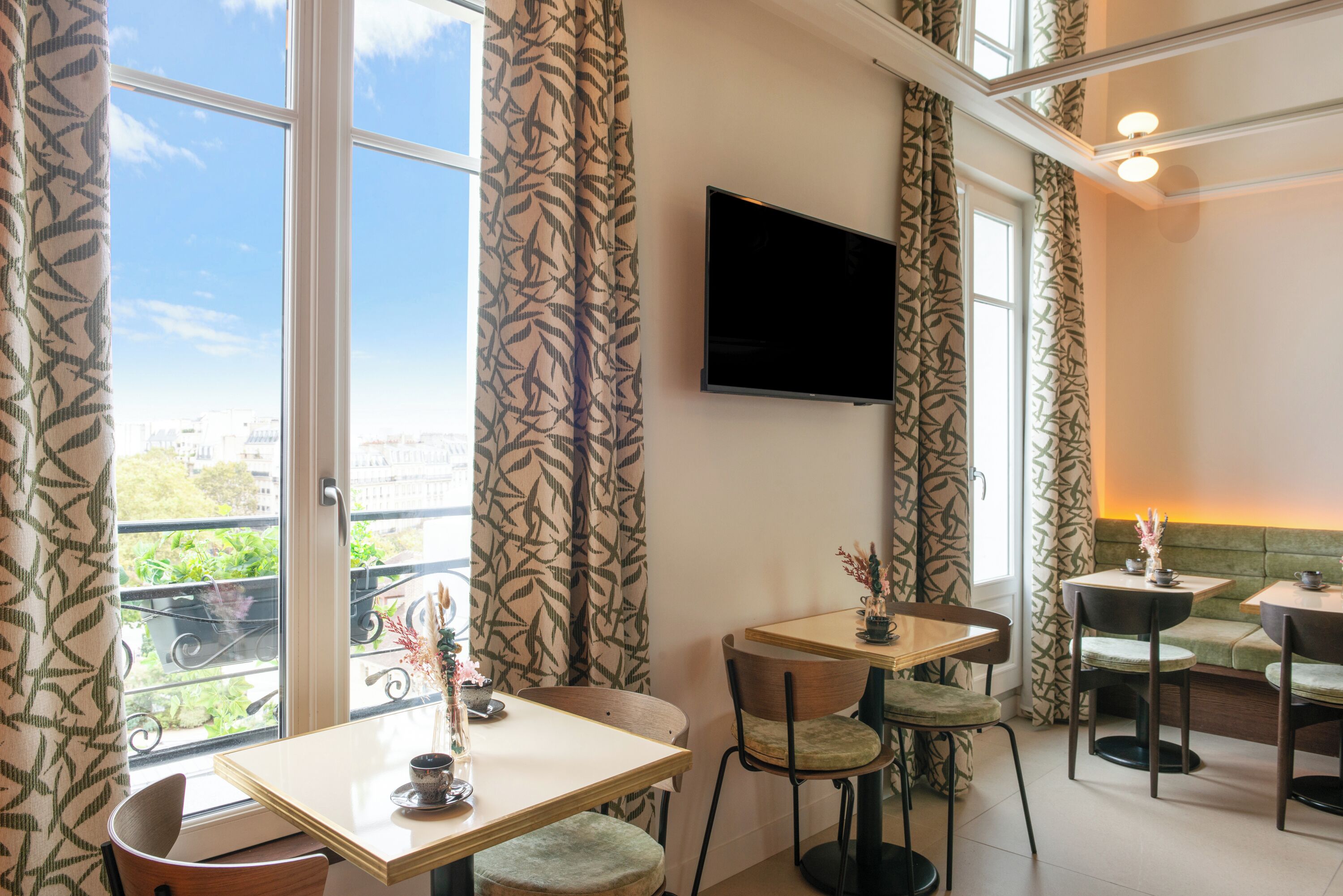 A dining area with a large window, a flat-screen TV mounted on the wall, and a balcony with a view of greenery.
