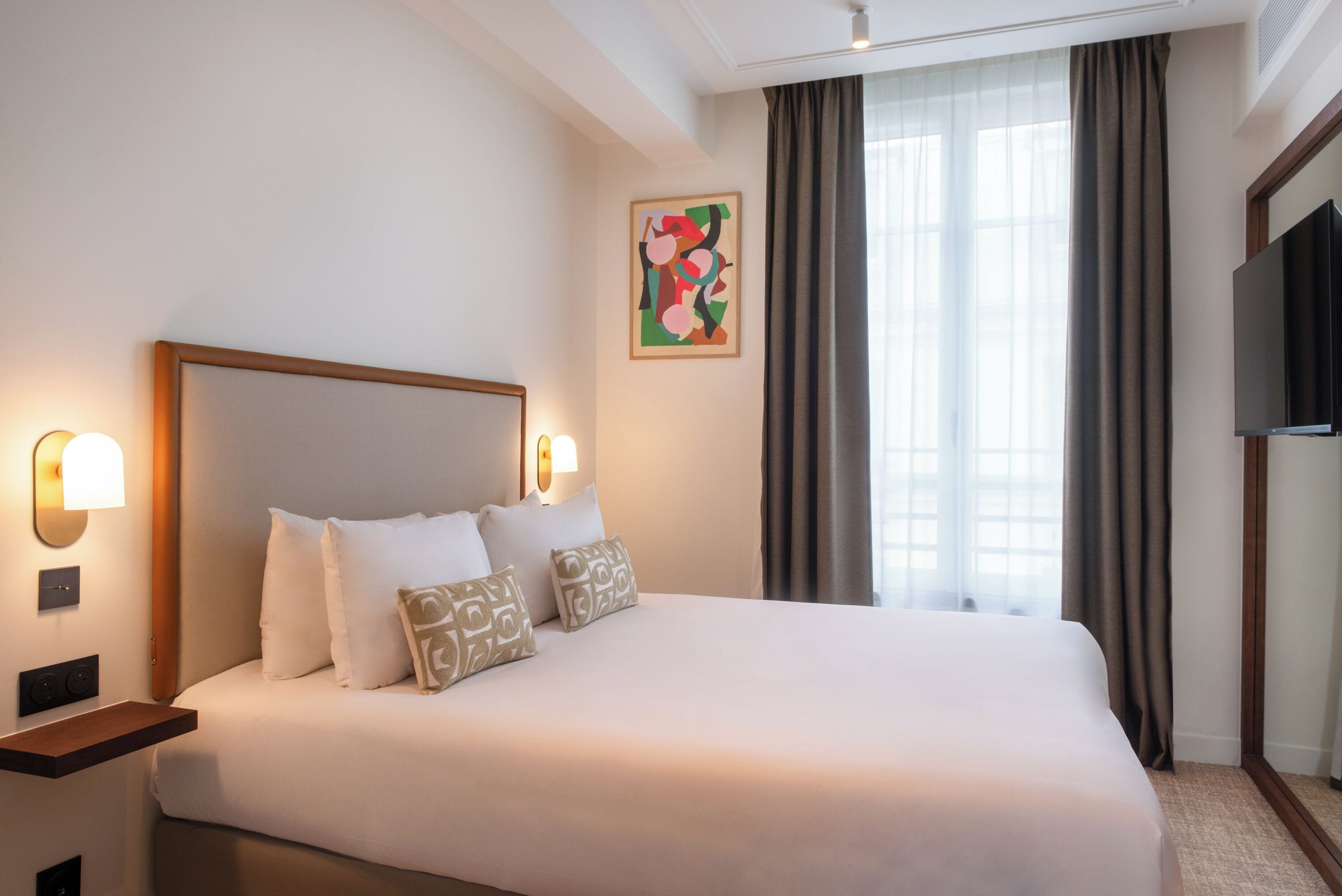 A hotel room with a large bed, two bedside tables, a flat-screen TV, and a window with curtains.