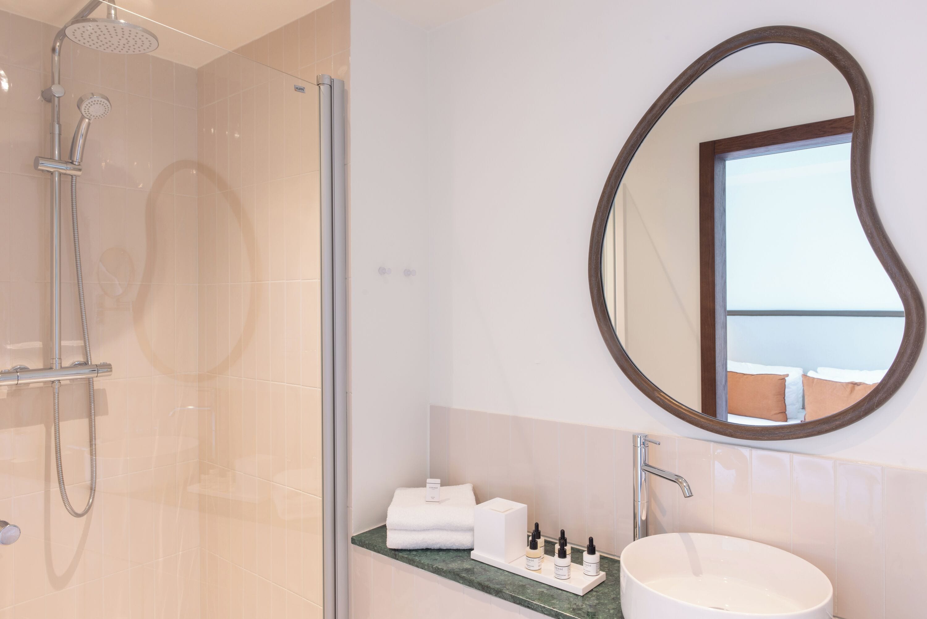 Baño con ducha envuelta en vidrio, espejo redondo, lavamanos y artículos de aseo en la encimera.