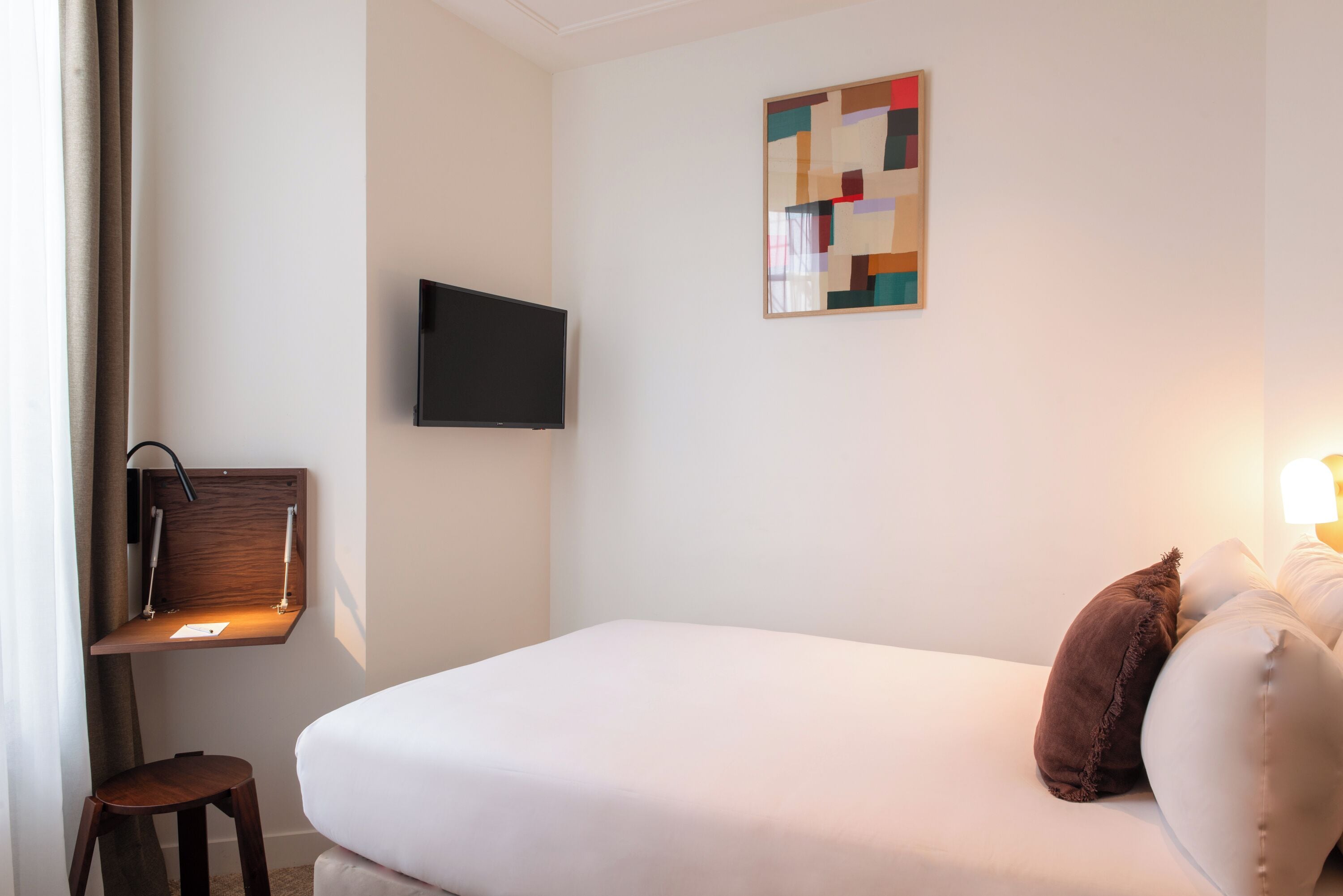 A hotel room with a bed, a mounted TV, a small desk with a chair, and a wall-mounted artwork.