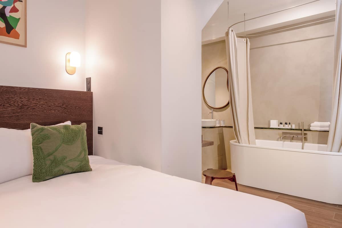 Une chambre d’hôtel avec un grand lit, une tête de lit en bois, un oreiller vert, une lampe fixée au mur, une salle de bain avec une baignoire, un miroir rond et un petit tabouret en bois.