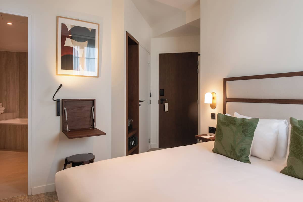 Une chambre d’hôtel avec un lit, une table de chevet, une étagère fixée au mur, une porte et un tableau encadré accroché au mur.