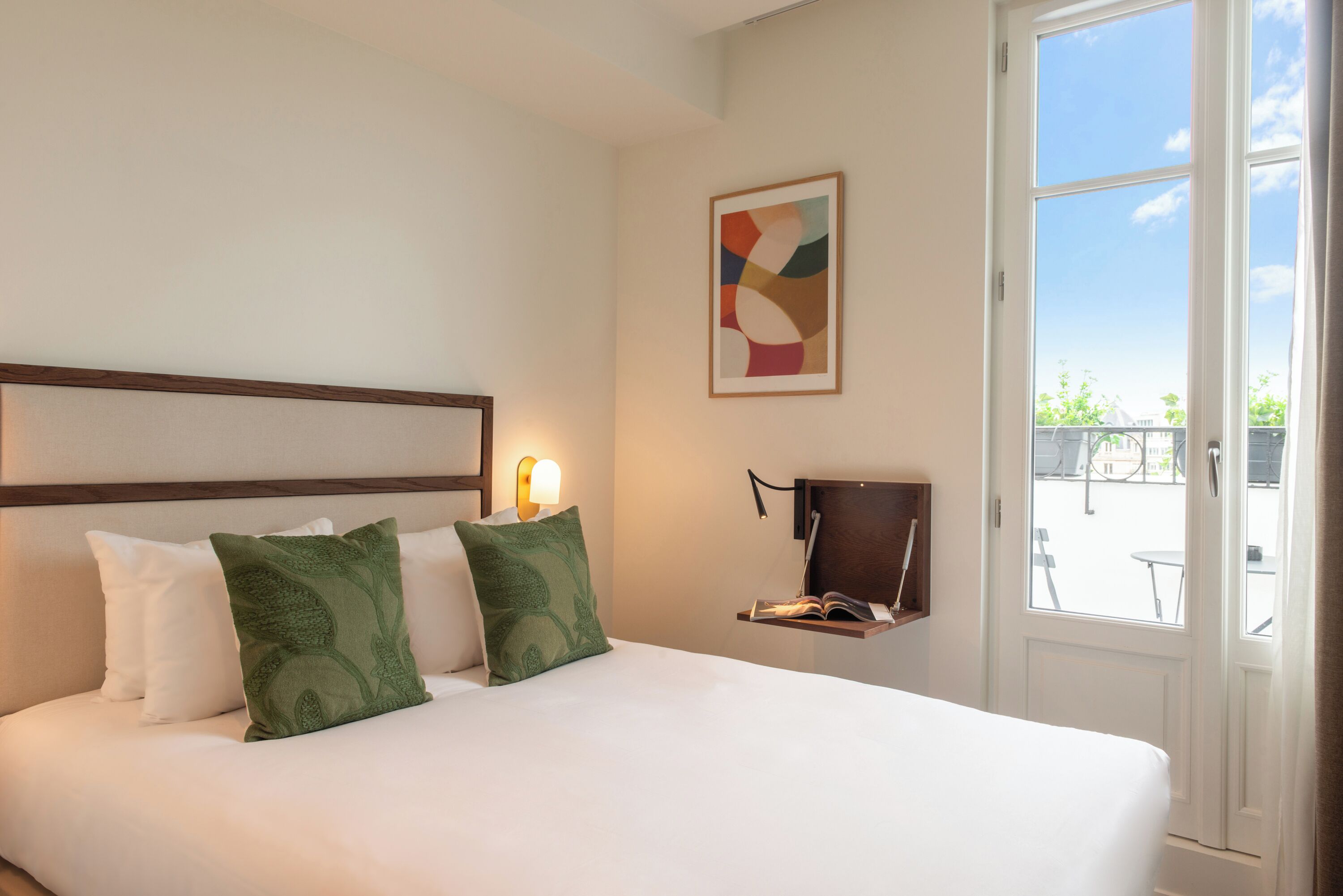 A hotel room with a bed, pillows, a bedside table with a lamp, a framed abstract painting, and a view through a door.