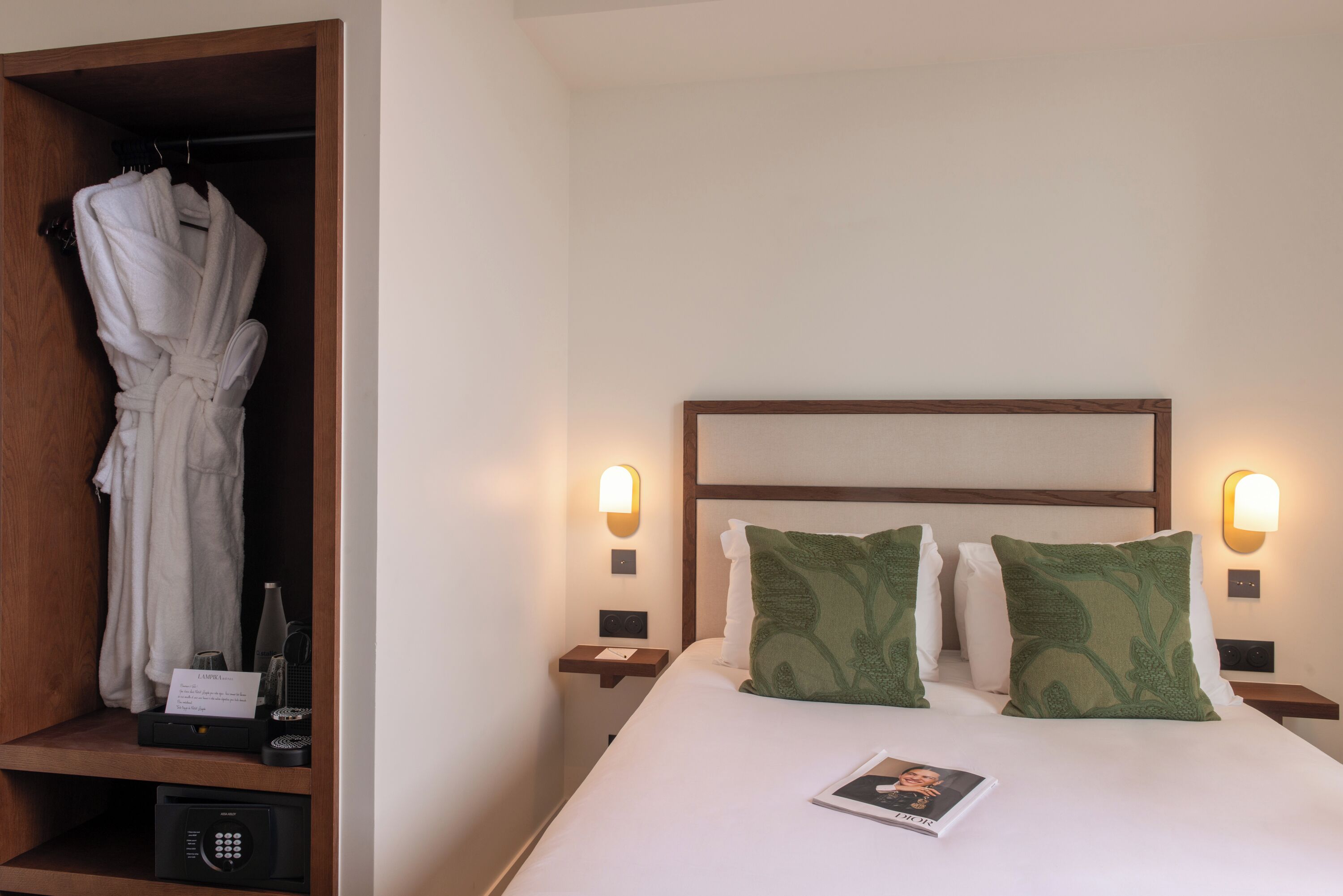 A hotel room with a bed, two pillows, a robe hanging, a nightstand, a bedside table, and a wall-mounted light fixture.