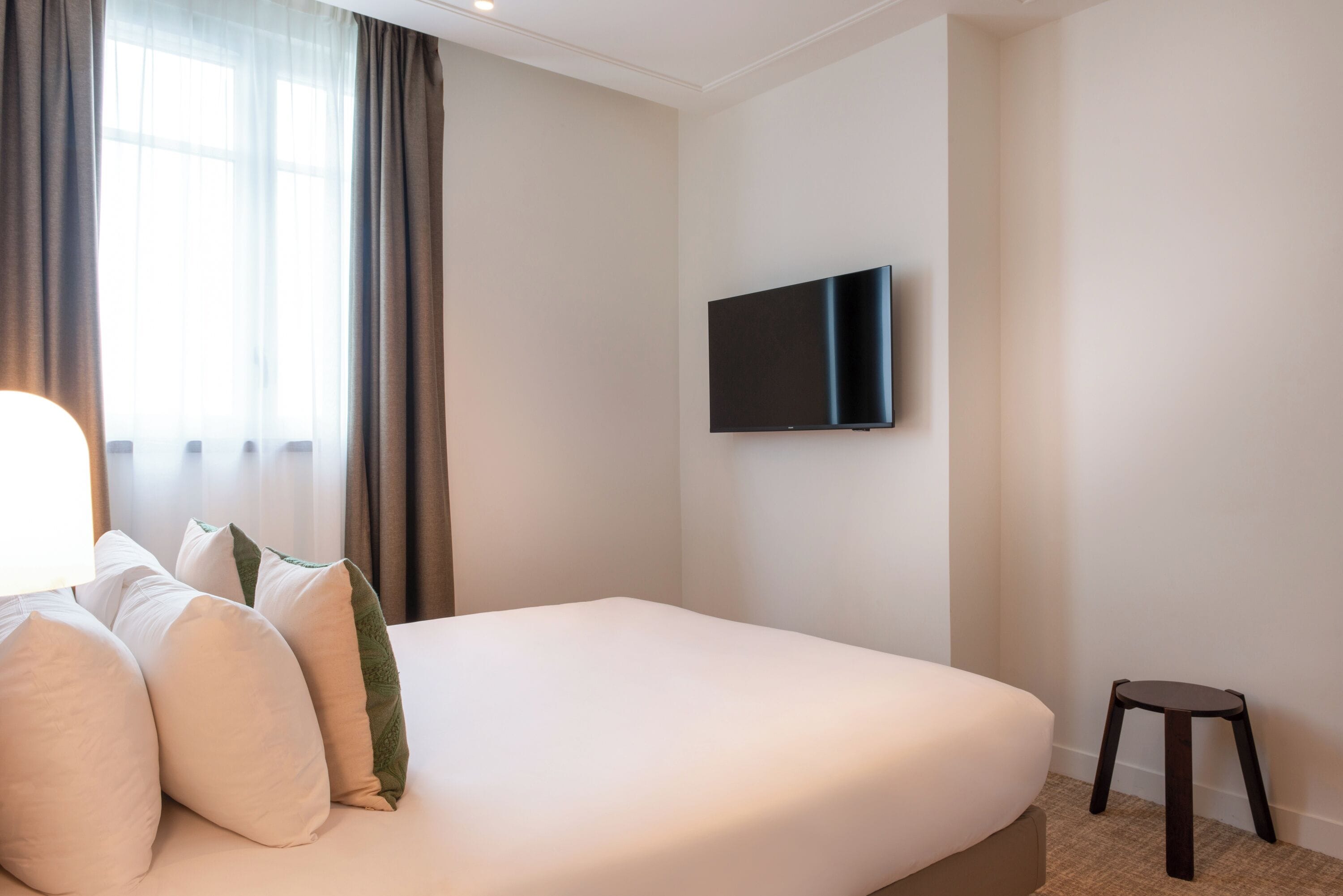 Habitación de hotel con una cama grande, un televisor de pantalla plana montado en la pared, un taburete pequeño y una ventana con cortinas.