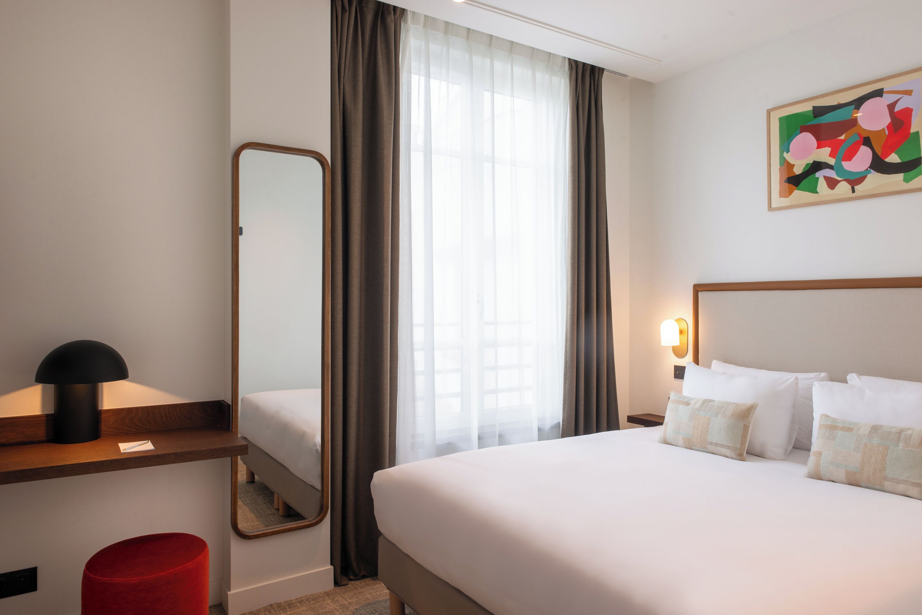 A hotel room with a bed, a bedside table, a mirror, a red ottoman, a window with sheer curtains, and a wall-mounted light fixture.