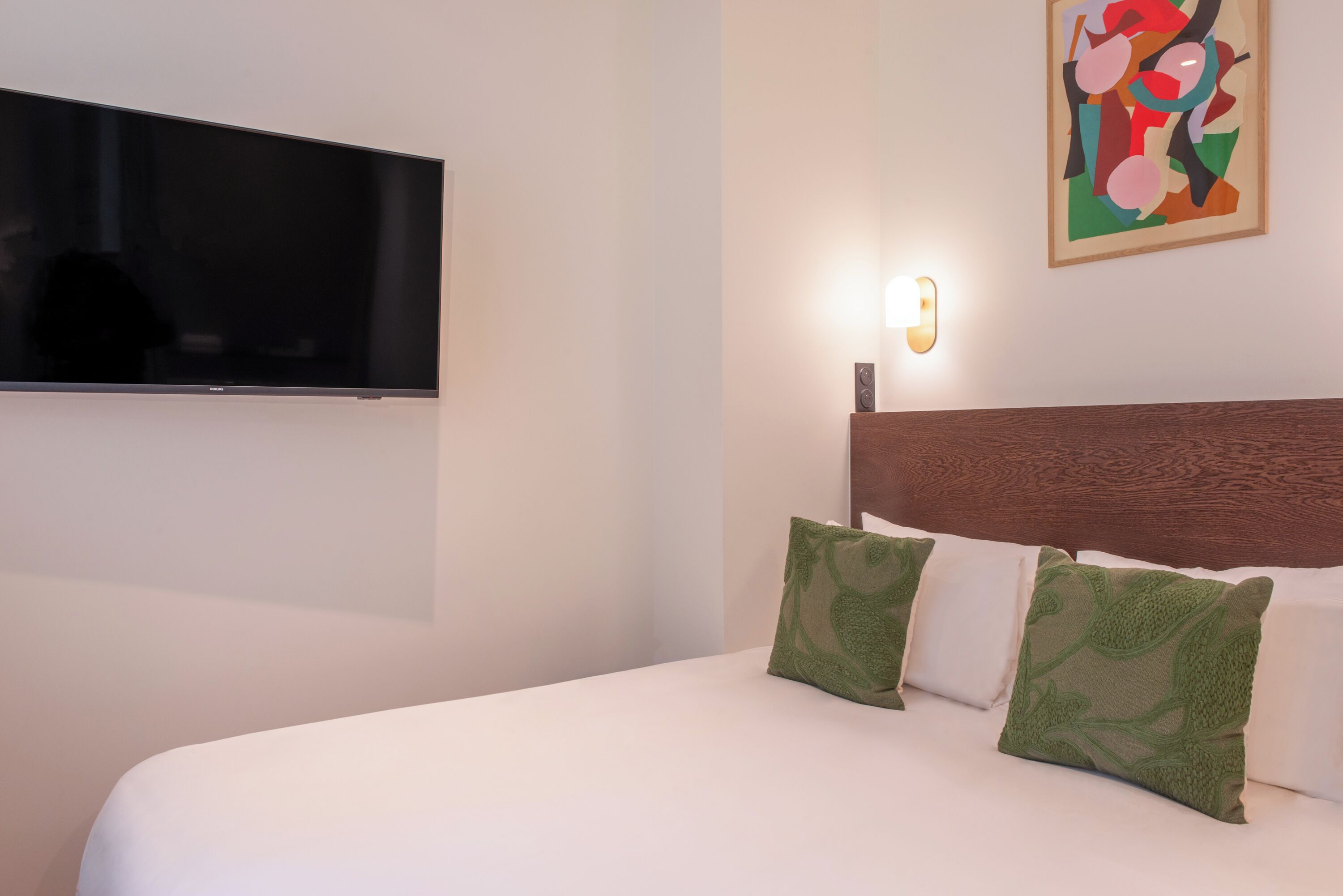 A hotel room with a flat-screen TV mounted on the wall, a bed with white bedding and green patterned pillows, a wooden headboard, and a colorful abstract painting on the wall.