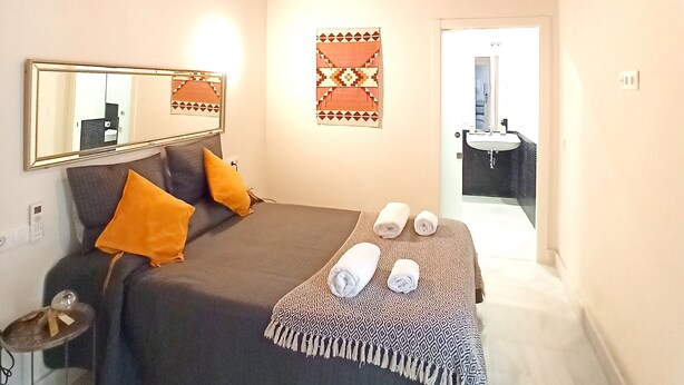 Une chambre à coucher comprenant un lit, une table de chevet, un miroir, un climatiseur fixé au mur et une porte donnant sur une salle de bain.