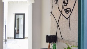 Un couloir avec un luminaire suspendu au plafond, fait de fibres tressées, une tapisserie représentant un visage, une console en bois sur laquelle est posée une plante, et un sol à motifs.