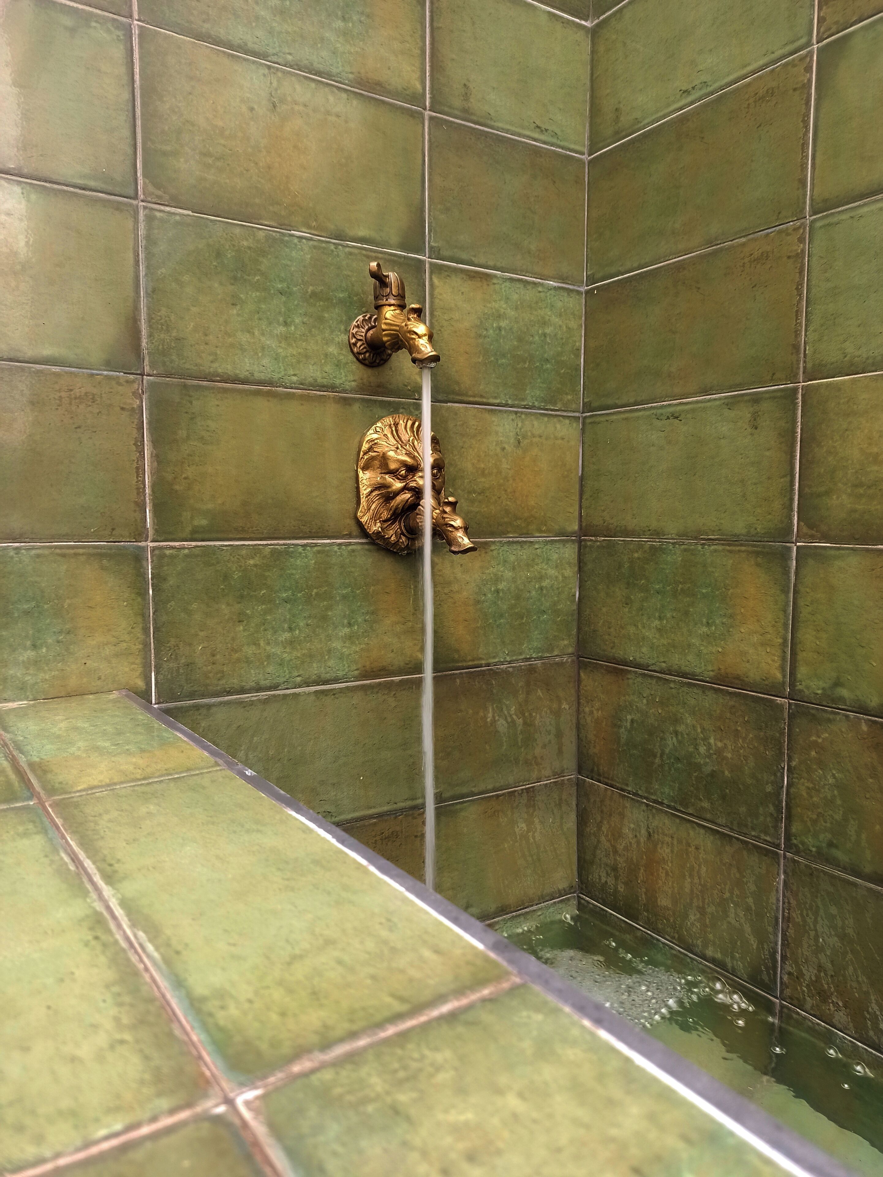 A shower with green tiles and two gold faucets.