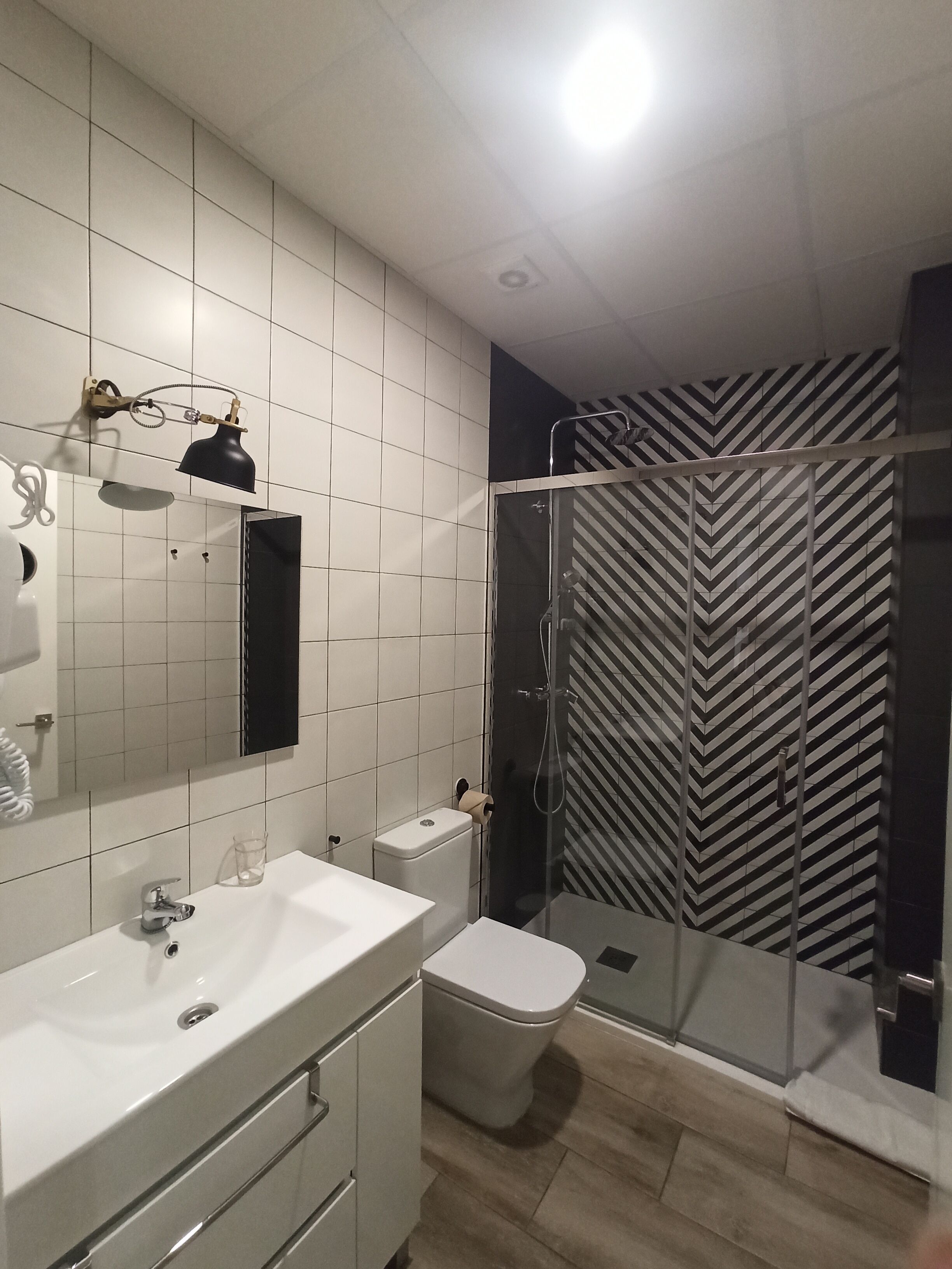 A modern bathroom with a white sink, mirror, and toilet. There is a glass shower enclosure with a black and white striped shower curtain.