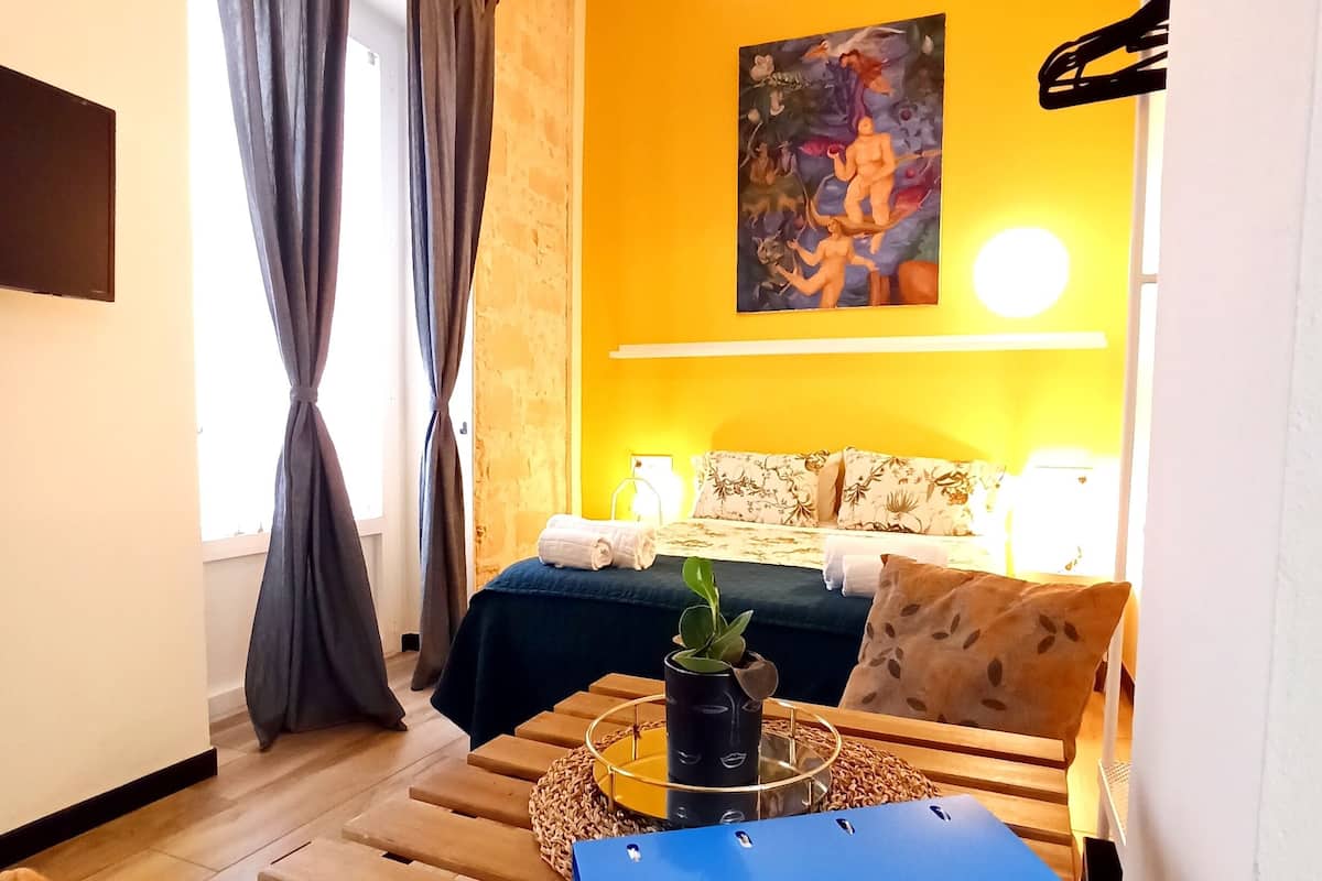 Een gezellige kamer met een bed, een bureau en een stoel. Er staat een plant op het bureau en een schilderij aan de muur.