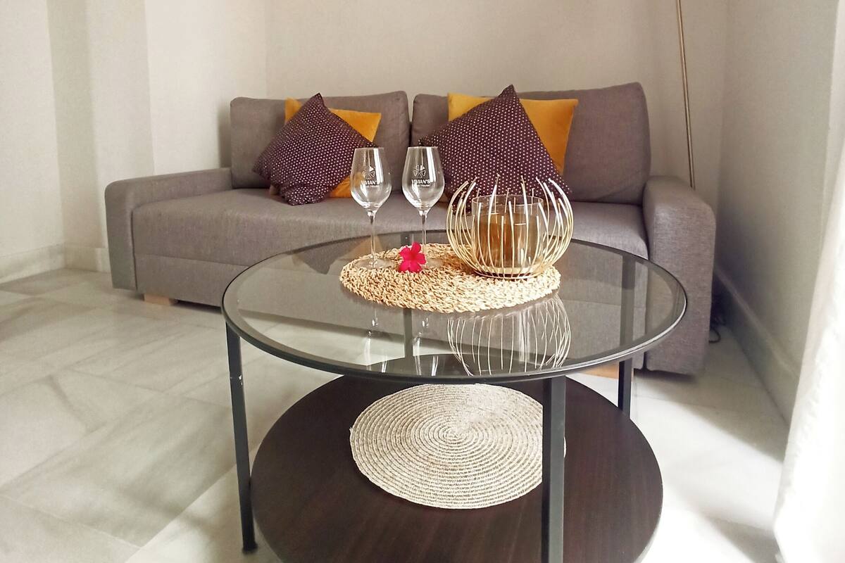 Een woonkamer met een ronde glazen salontafel, een bank met kussens, een vloerlamp en een spiegel aan de muur.