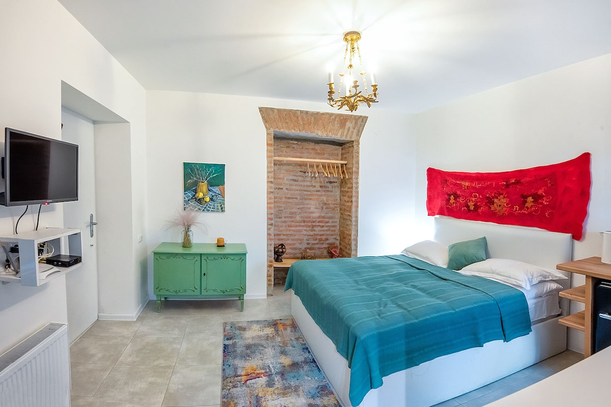 Quarto com cama, televisão, penteadeira verde, tapeçaria vermelha e lustre.