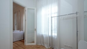 A bedroom with a wooden floor, a white bed, a door leading to another room, and a metal drying rack against the wall. - HB apartments Tabukashvili (Tbilisi)