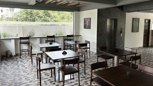 A modern dining area with wooden tables and chairs, large windows with a view of greenery, and a stone accent wall. - Palladium Luxury Suites Unit II (Hyderabad)