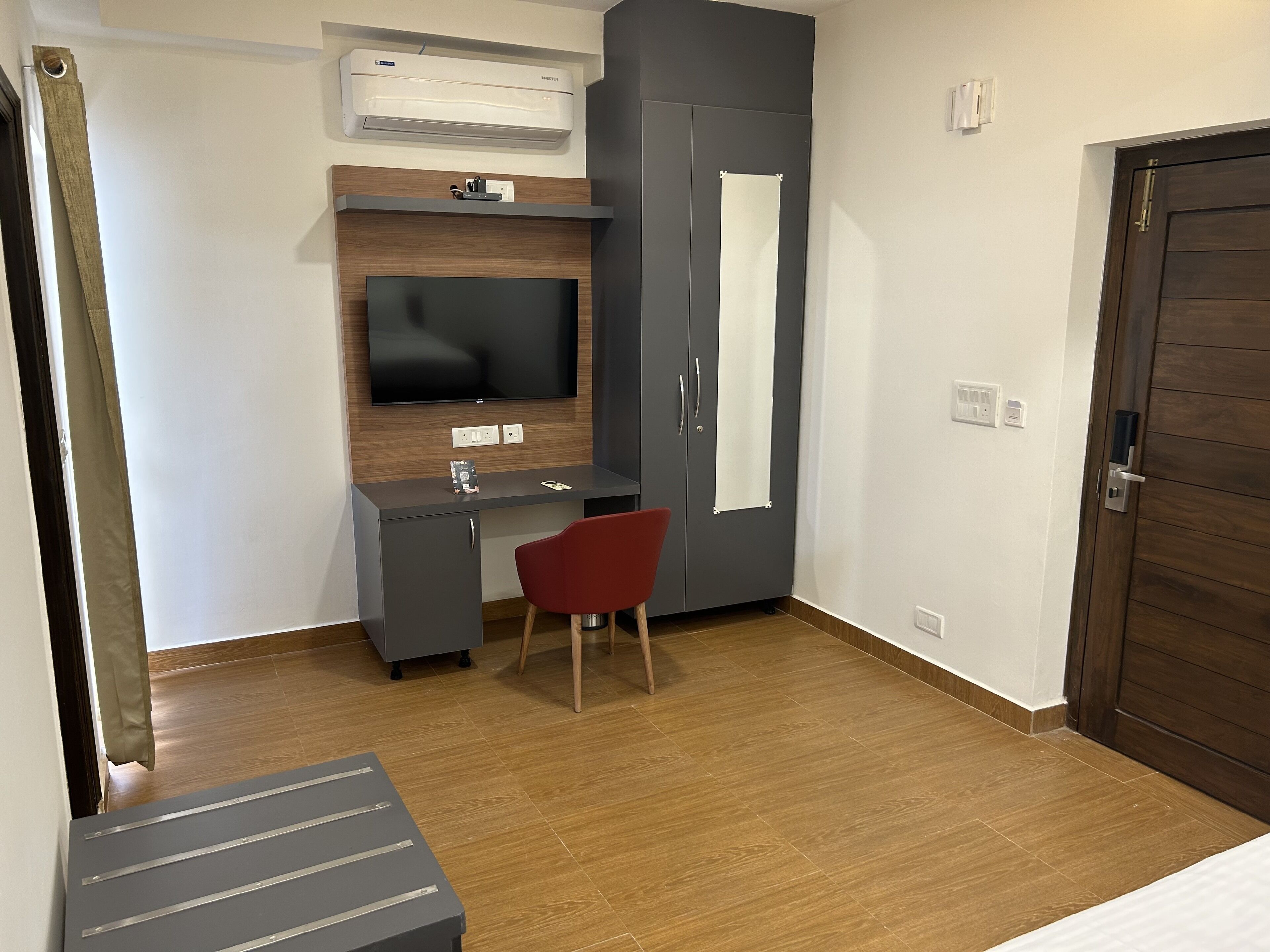 A compact room with a grey desk, a red chair, a flat-screen TV mounted on a wooden panel, a wardrobe with a mirror, and a door with a handle.