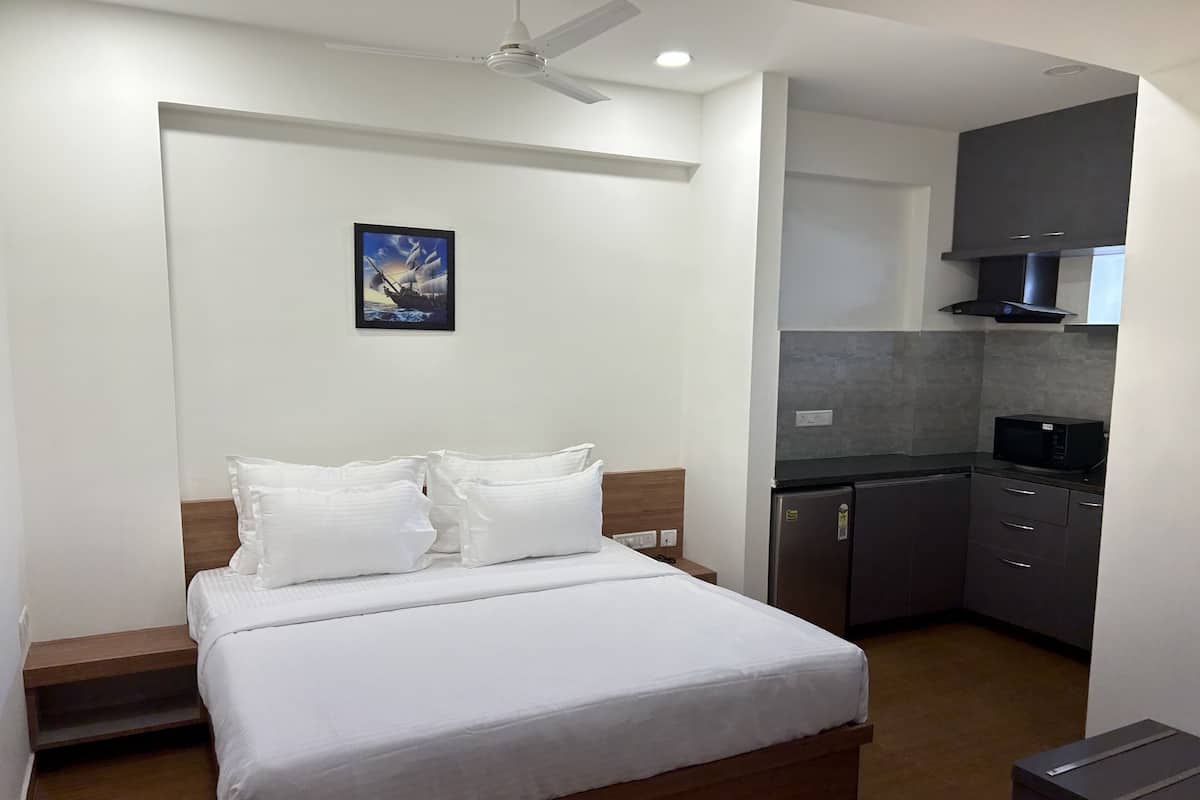 A bedroom with a bed, pillows, a nightstand, a ceiling fan, and a small kitchen area with a microwave and cabinets.