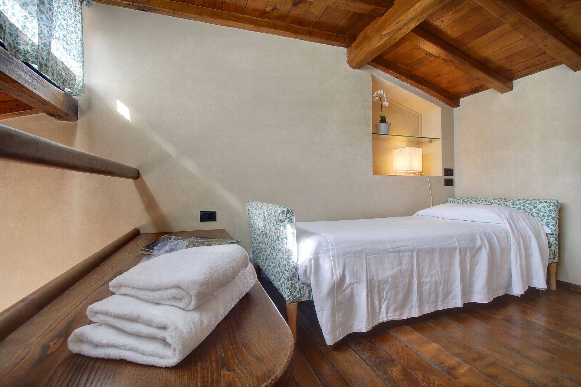 Ein Schlafzimmer mit Holzboden, einem Bett mit weißer Bettwäsche, einem kleinen Nachttisch und einem Fenster mit Vorhang.