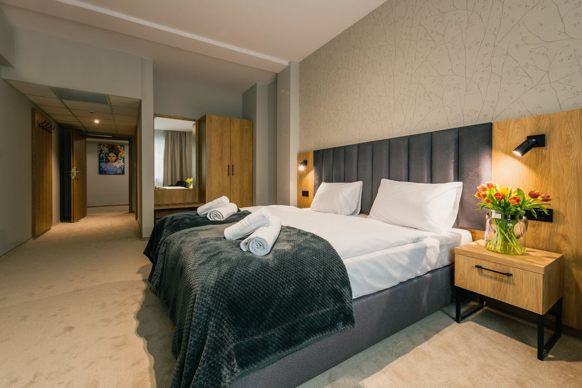 A modern hotel room with a large bed, a wooden wardrobe, a bedside table with a vase of flowers, and a mirror above the bed.