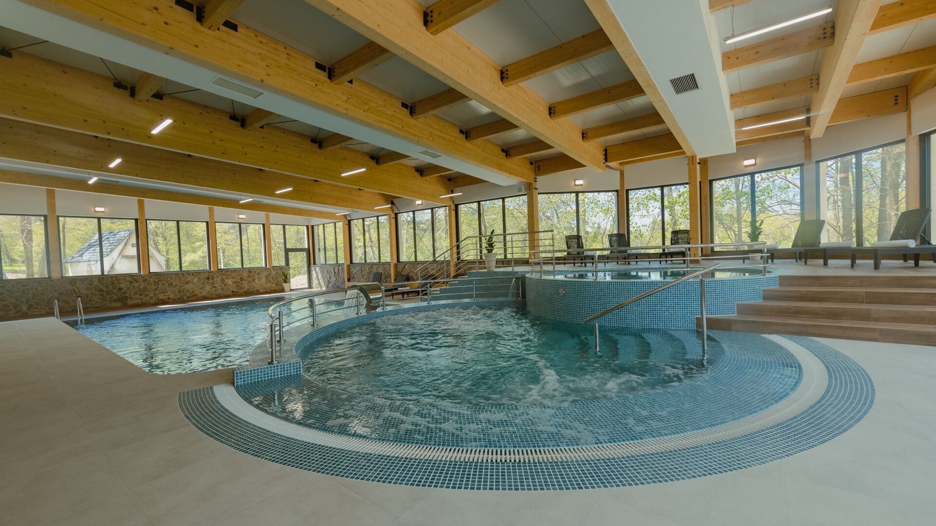 An indoor swimming pool with a tiled edge, wooden beams, and large windows.