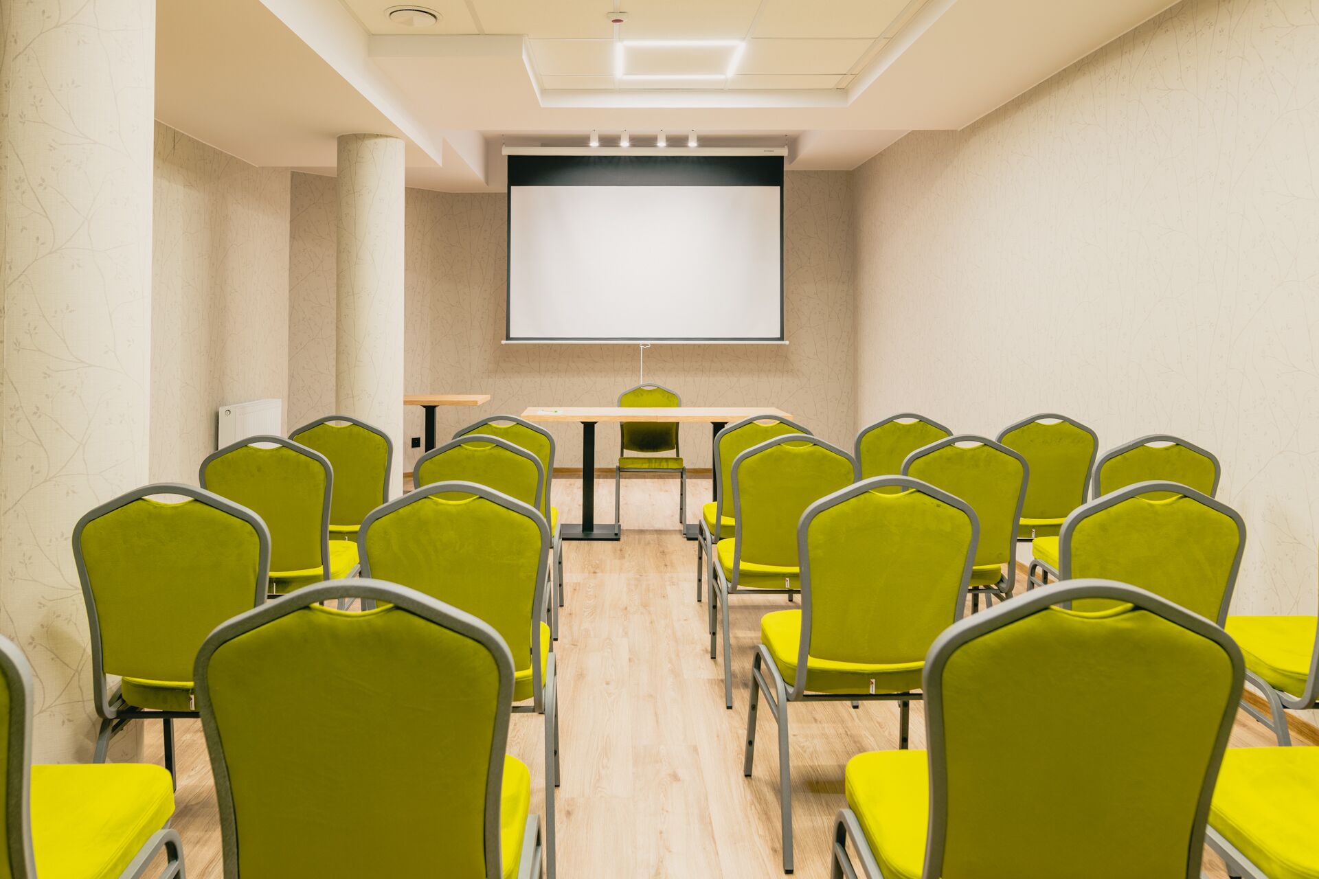 Un salón de conferencias con una pantalla grande, varias filas de sillas amarillas y una mesa de madera.