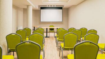Un salón de conferencias con una pantalla grande, varias filas de sillas amarillas y una mesa de madera.