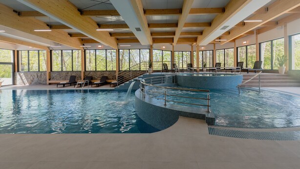 Una piscina interior con suelo de baldosas, vigas de madera y grandes ventanales.