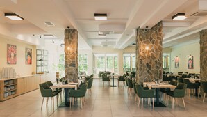 Un restaurante espacioso con techos altos, pilares de piedra y ventanas amplias. Las mesas están dispuestas en forma de U, y el suelo está cubierto de baldosas.