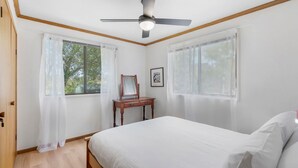 Un dormitorio con cama, ventilador de techo, cómoda y dos ventanas con cortinas.