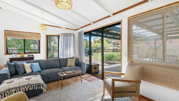 Un salón amplio con un sofá gris, una mesa de centro de madera y un sillón beige. Hay un amplio ventanal corredero que da a un balcón con vistas a zonas verdes.