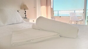 A neatly made bed with folded white linens, a bedside lamp, a chair by the window, and a view of the sea.