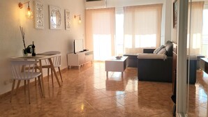 A modern living room with a sofa, coffee table, and dining area with chairs. There are wall-mounted lights and a ceiling light fixture.