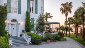 A white building with green shutters, a covered outdoor seating area, and a paved pathway leading to the entrance. - La Villa Mauresque (Saint-Raphaël)