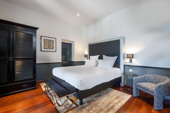 A modern bedroom with a large bed, a dark wooden wardrobe, a small round table, and a patterned armchair.