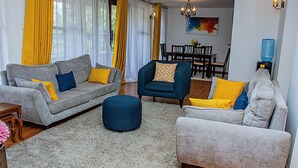 Ein Wohnzimmer mit einem grauen Sofa, einem blauen Sessel und einem grauen Sessel mit gelben Kissen. In der Mitte steht ein blauer Hocker.