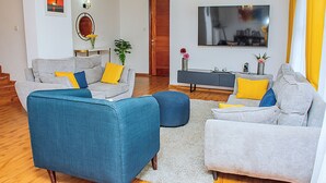 Ein Wohnzimmer mit zwei grauen Sofas, eines mit blauen und gelben Kissen, und einem blauen Sessel. Ein weißer Teppich, Holzboden, ein Fernseher an der Wand und eine Tür im Hintergrund.