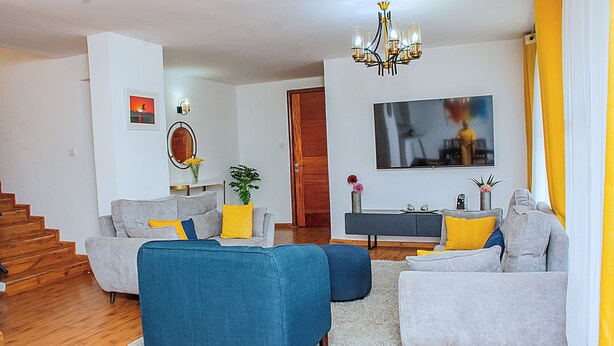 Ein modernes Wohnzimmer mit Treppe, grauen Sofas mit blauen und gelben Kissen, Holzboden und einem großen Fernseher an der Wand.