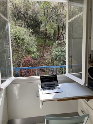 Habitación con un ventanal grande que da a una zona boscosa, una computadora portátil sobre un escritorio y una silla.