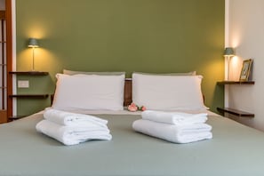 Una cama bien hecha con almohadas blancas y toallas dobladas, dos lámparas de noche y una pequeña estantería con un marco de fotos.