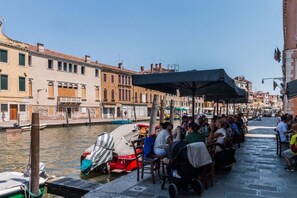 A canal-side restaurant with outdoor seating, a row of buildings, and boats docked along the water. - Colombina House close to @S.Lucia Train Station (Venezia)