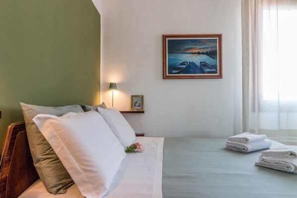 Una cama bien hecha con almohadas y toallas dobladas, un cuadro de una puesta de sol sobre el agua en la pared, y una lámpara en la mesita de noche.