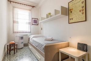 A bedroom with a bed, a desk, a radiator, a window with curtains, and a framed certificate on the wall.
