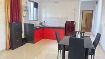 A compact kitchen with red cabinets, a microwave, and a refrigerator. There is a dining table with chairs and a ceiling fan.