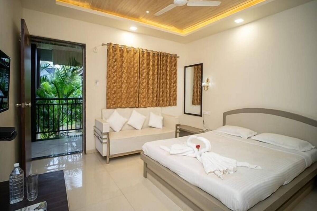 Habitación de hotel con cama, sofá, ventilador de techo, espejo y vista a un balcón con plantas.