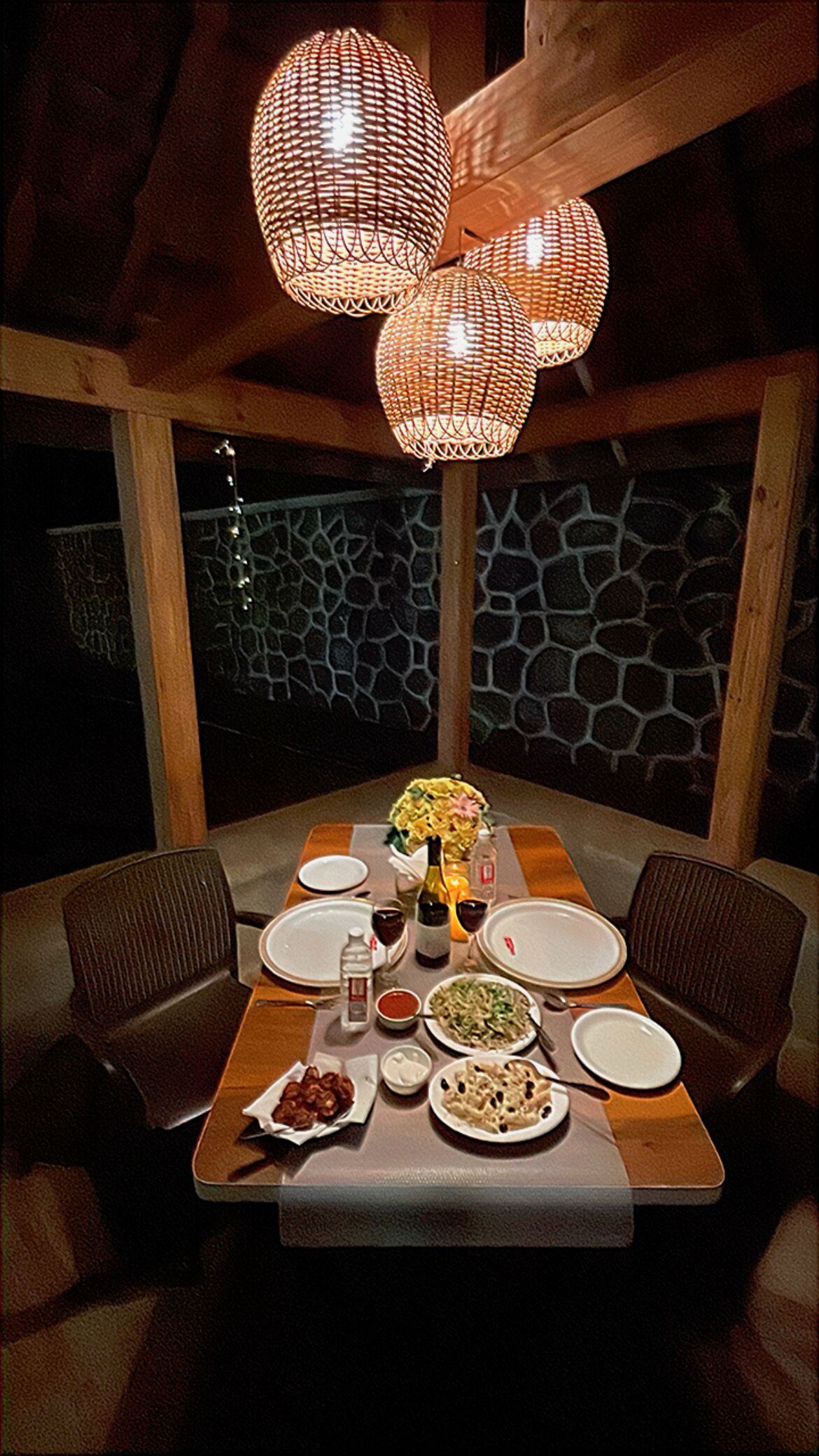 A dining table set for four with a stone wall backdrop, wicker chairs, and a woven light fixture above.